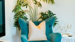 A teal armchair with a beige pillow, a small white side table with a gold 'LOVE' sign, and a large green plant with variegated leaves. - Peacock Palace (Springfield)