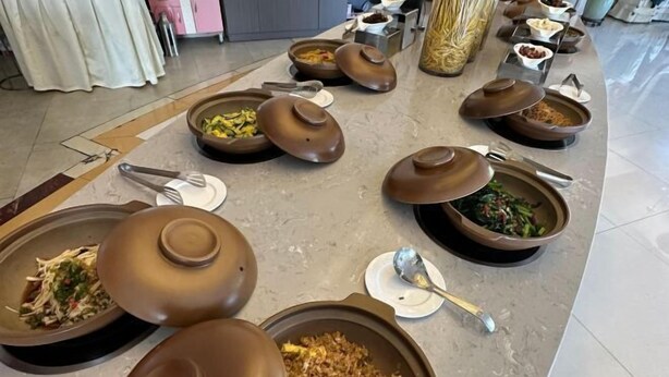 A buffet setup with various dishes in large, round, brown containers, some with lids, and serving utensils placed on the table.