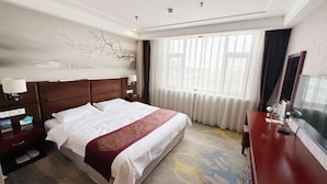 Une chambre d’hôtel avec un grand lit, une tête de lit en bois, une table de chevet avec une lampe, un bureau avec une chaise et une grande fenêtre avec des rideaux.