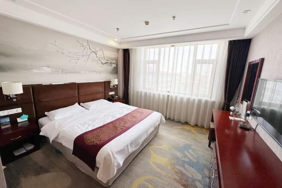 A hotel room with a large bed, a wooden headboard, a nightstand with a lamp, a desk with a chair, and a large window with curtains.