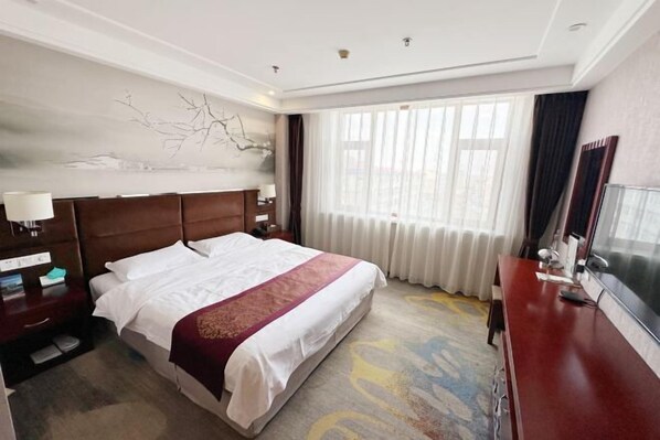 Une chambre d’hôtel avec un grand lit, une tête de lit en bois, une table de chevet avec une lampe, un bureau avec une chaise et une grande fenêtre avec des rideaux.