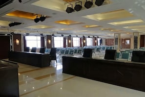 Une salle de conférence avec des rangées de chaises, une scène éclairée et de grandes fenêtres.