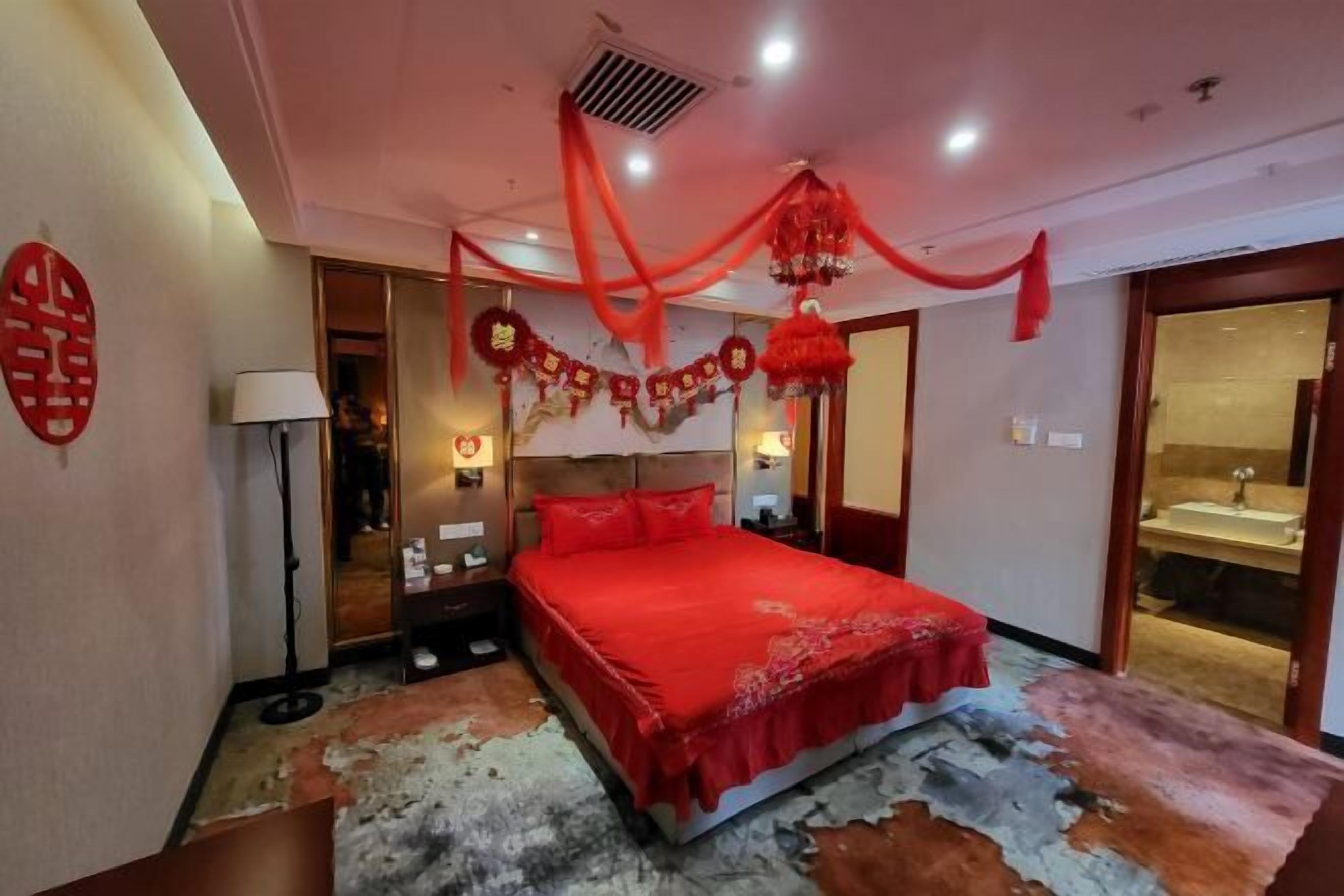 A bedroom with a red bedspread, a headboard, a bedside table, a lamp, and a wall decoration.