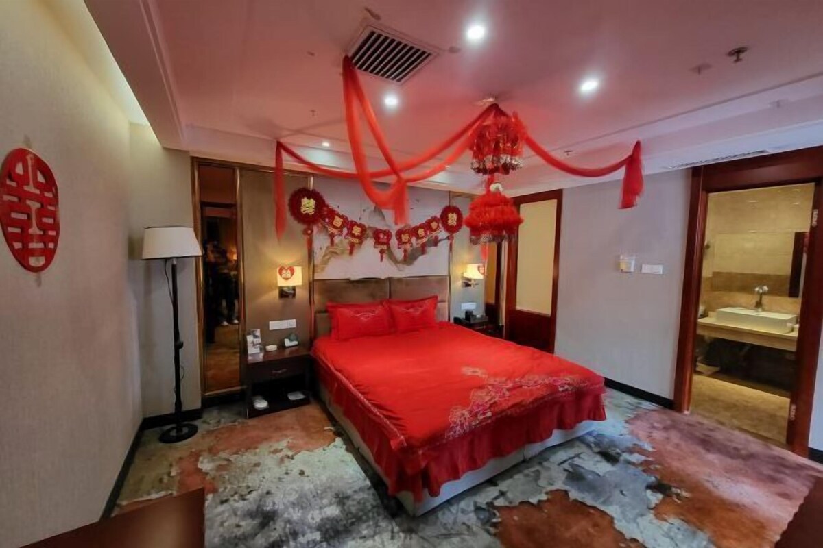 Une chambre à coucher avec une couvre-lit rouge, une tête de lit, une table de chevet, une lampe et un élément de décoration murale.