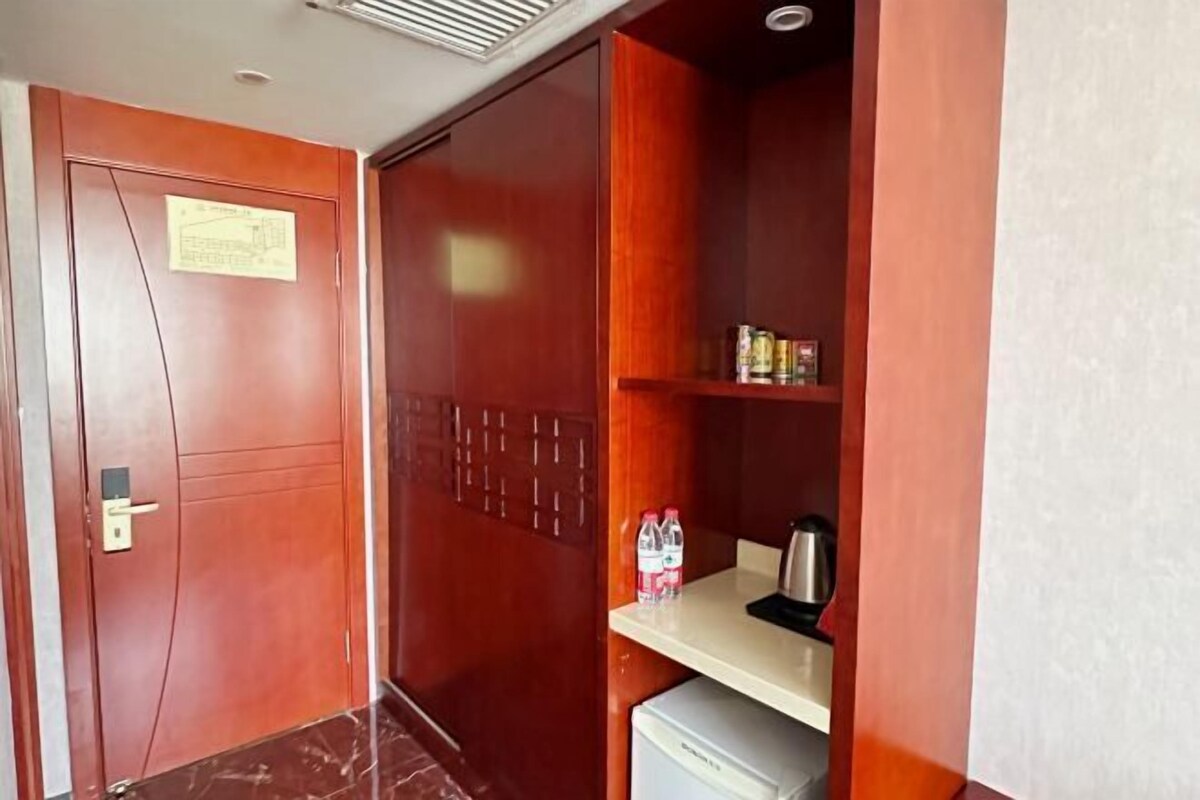 Une pièce de taille modeste avec une porte en bois, une petite kitchenette équipée d’un réfrigérateur, et une étagère contenant des bouteilles d’eau et une bouilloire.
