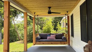 A covered porch with a wooden ceiling, a ceiling fan, a swing seat with cushions, and a stone sculpture. - Wow! Only 3 min to Gore Mtn! King bed too (North Creek)