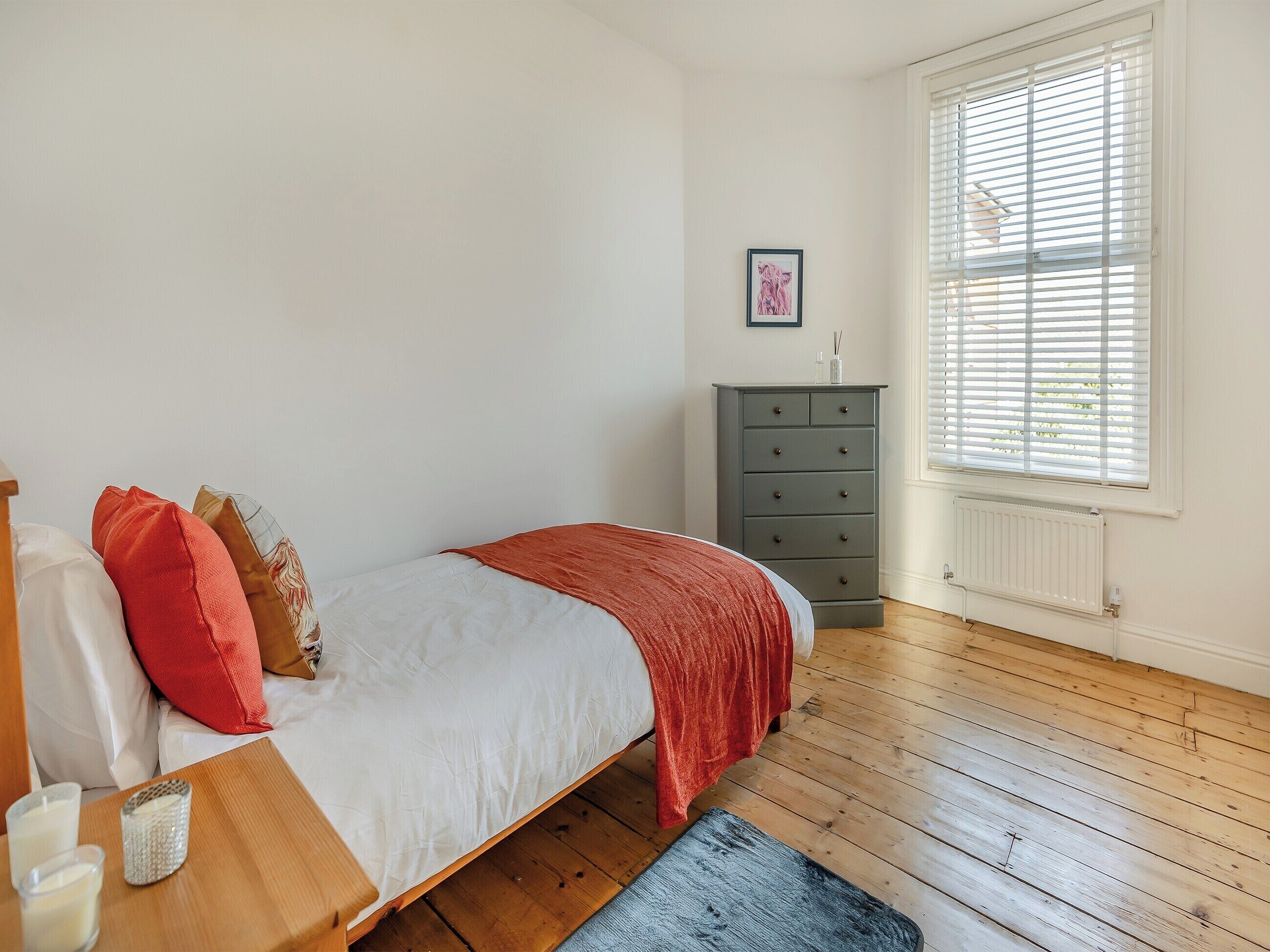 Een slaapkamer met een bed, een dressoir, een raam met zonwering en een radiator.