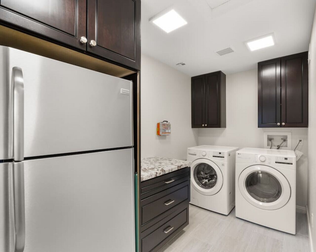 A compact kitchen with a stainless steel refrigerator, two white washing machines, dark wooden cabinets, and a marble countertop.