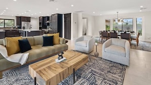 A spacious living room with a sofa, armchairs, and a coffee table. There is a dining area with a table and chairs, and a kitchen in the background.