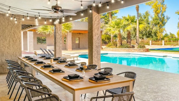 An outdoor dining area with a pool view, featuring a long table set for dining, lounge chairs, and string lights.