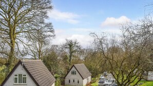 A rural setting with two houses, one with a sloped roof and green shutters, and another with a flat roof. There are cars parked on the side of the road, and a tree in the foreground.