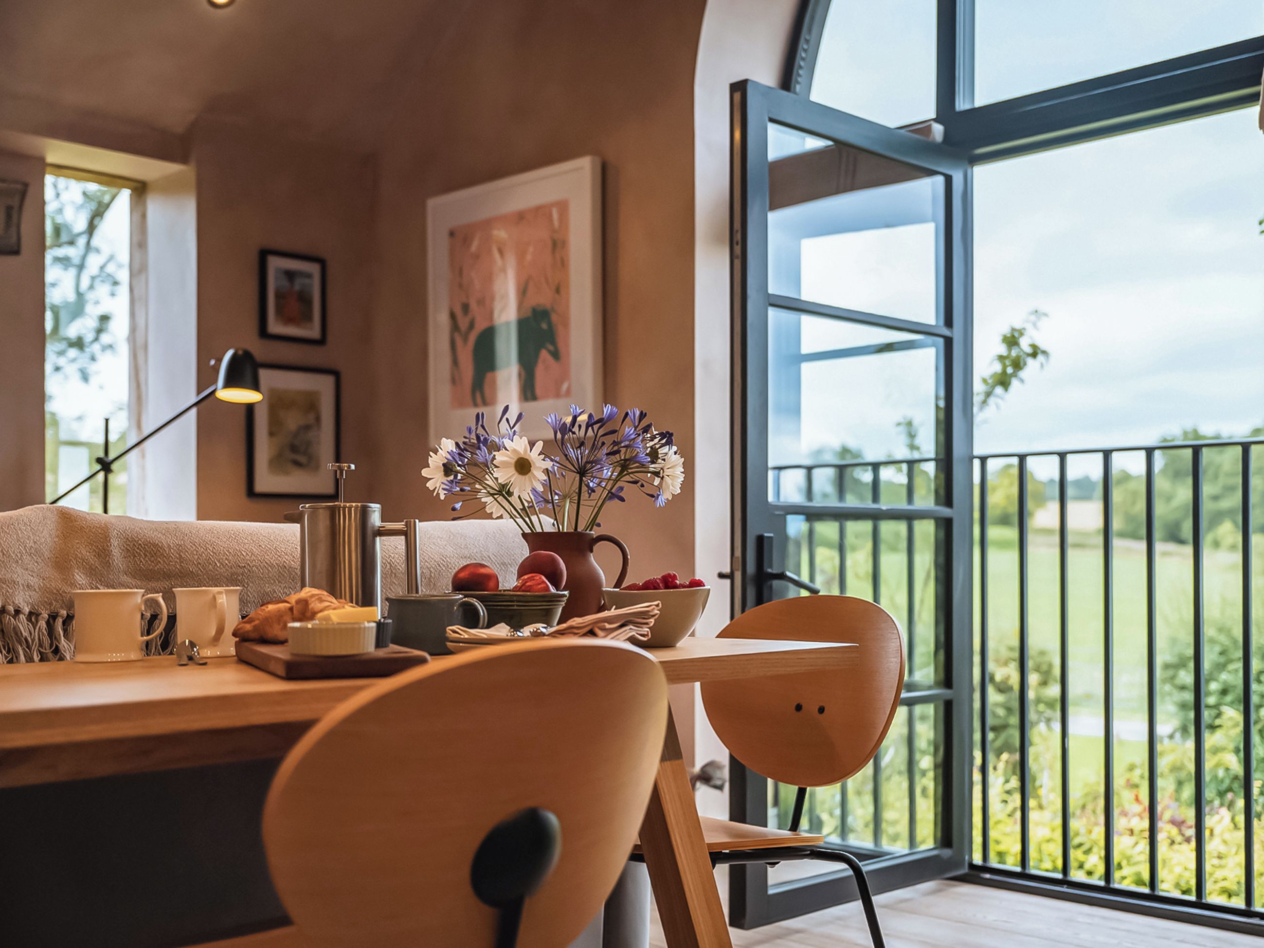 A modern dining area with a wooden table, chairs, and a vase of flowers. A large window offers a view of greenery outside.