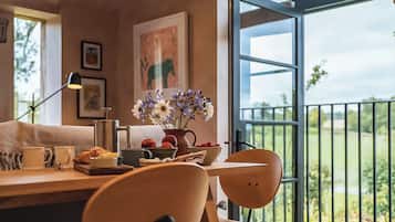 A modern dining area with a wooden table, chairs, and a vase of flowers. A large window offers a view of greenery outside.