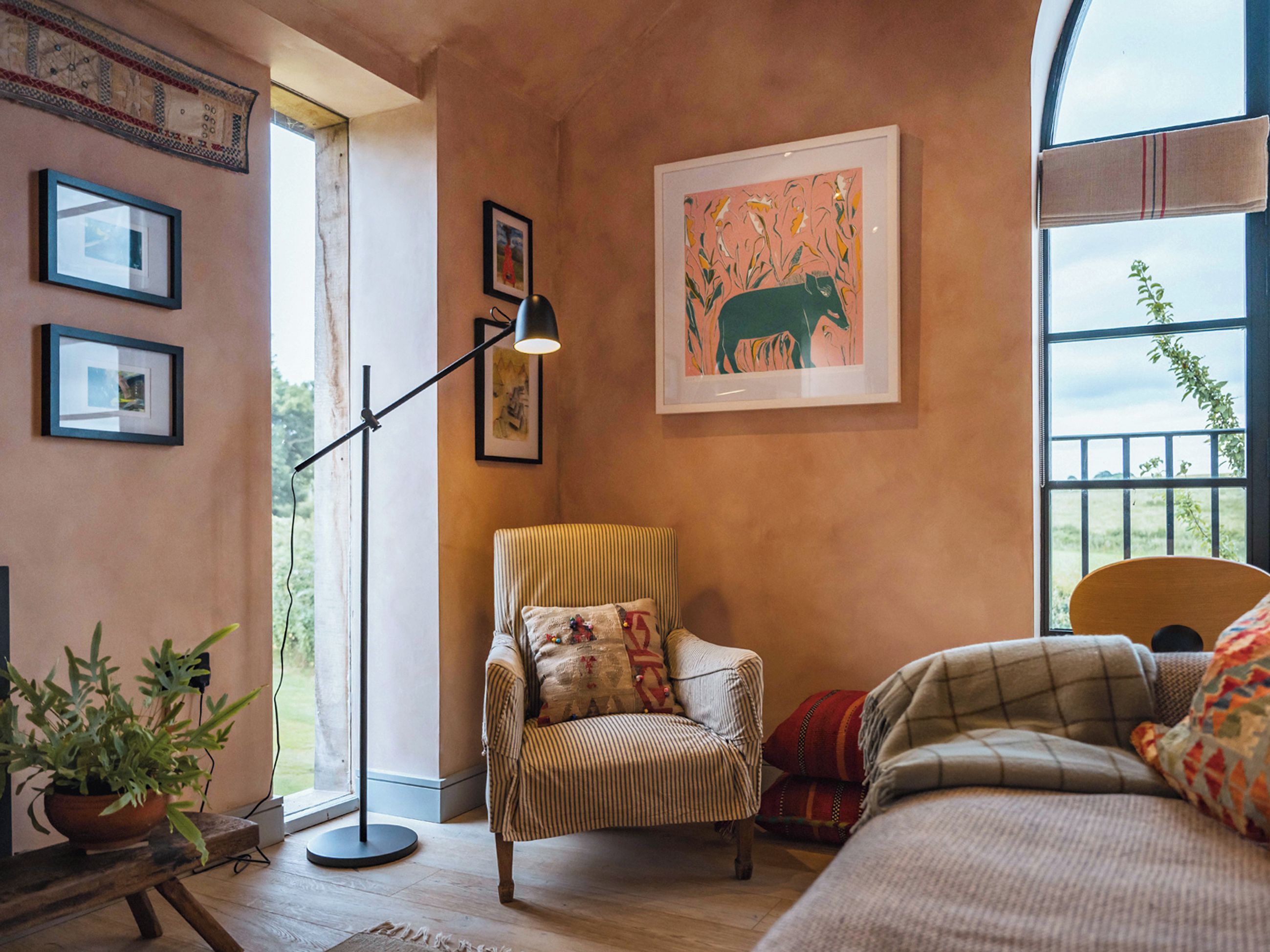 Une pièce chaleureuse avec un fauteuil à rayures, un canapé et une lampe sur pied. Les murs sont ornés de tableaux encadrés et une fenêtre offre une vue sur la verdure.