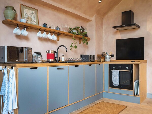 A compact kitchen with a microwave, toaster, kettle, and oven. There are hanging cups and a shelf with decorative items. - 1 bedroom accommodation in Willington, near Tarporley (Willington, near Tarporley)