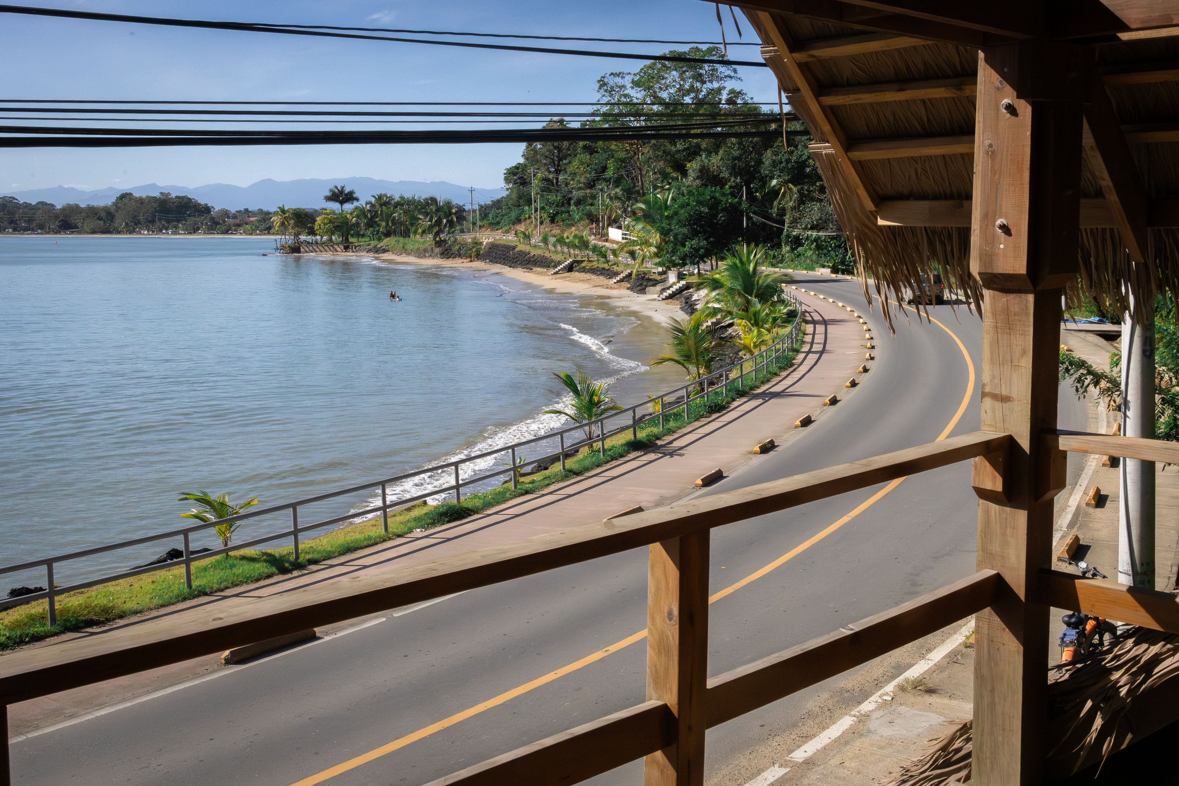 A coastal road with a wooden bridge, palm trees, and a view of the sea and mountains.
