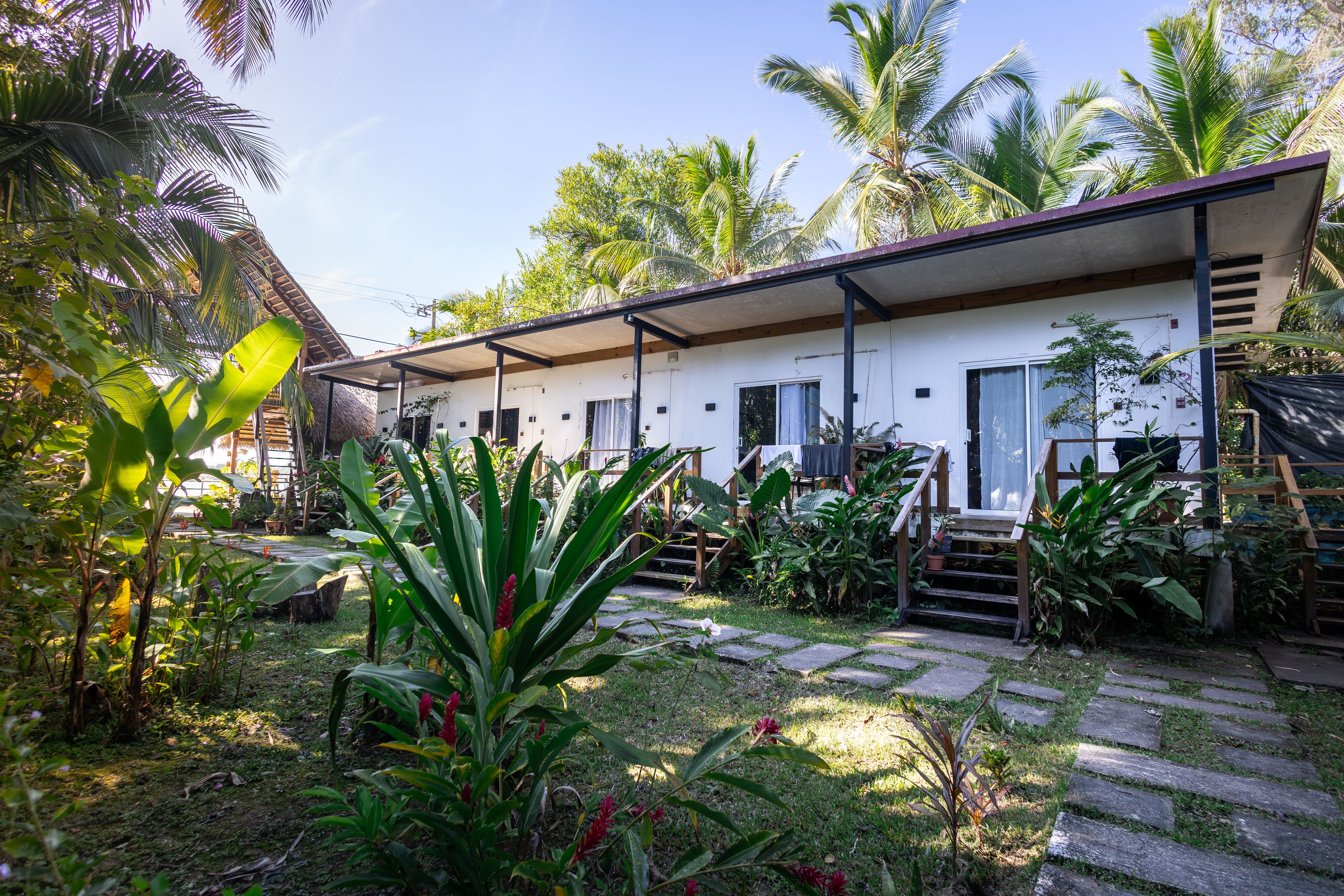 Um imóvel residencial com varanda coberta, escadaria que leva à entrada e jardim com plantas tropicais.