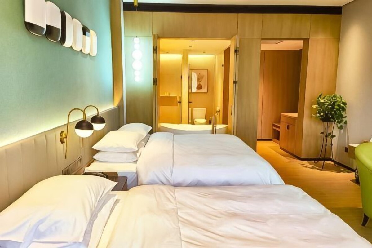 A hotel room with two beds, a green chair, a plant, and a bathroom visible through an open door.