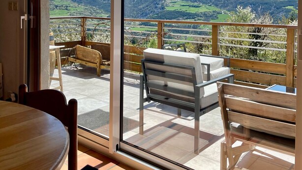 A balcony with a table and chairs, offering a view of a mountain and valley.