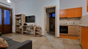 Un salon moderne avec un canapé gris, des étagères en bois ornées de plantes, un téléviseur à écran plat et une cuisine avec des meubles orange et un four encastré.