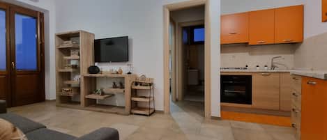 A modern living room with a grey sofa, wooden shelving with plants, a flat-screen TV, and a kitchen with orange cabinets and a built-in oven.