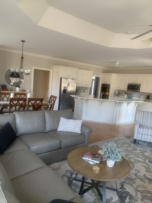 A modern living room with a grey sofa, a coffee table with a magazine and a plant, and a dining area with wooden chairs and a chandelier. - Master’s Rental - Golf course view in Woodside’s beautiful gated community! (Aiken)