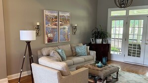 A living room with a beige sofa, a floor lamp, two wall-mounted paintings, and a chandelier. - Master’s Rental - Golf course view in Woodside’s beautiful gated community! (Aiken)