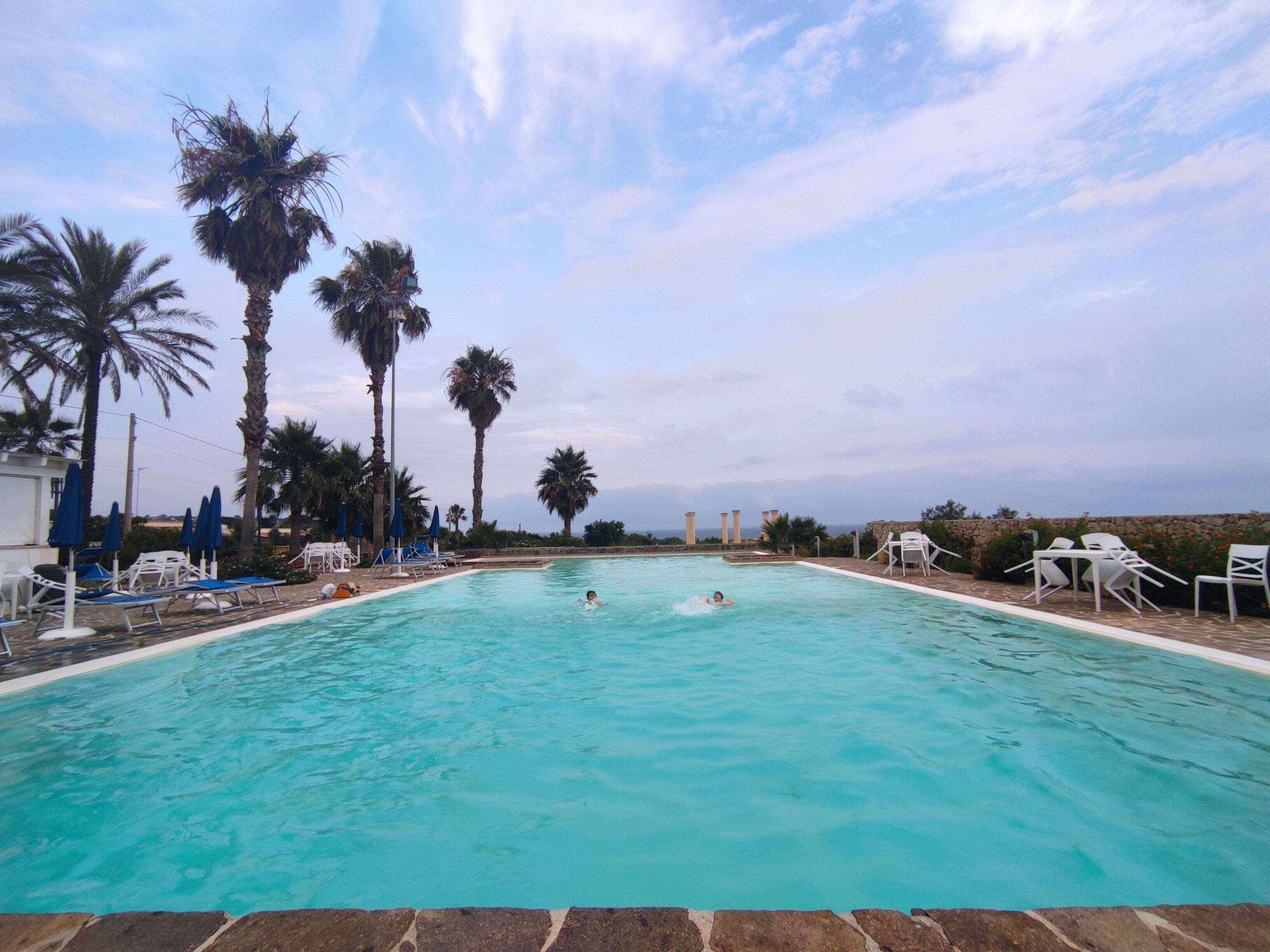A swimming pool with two people swimming, surrounded by palm trees and lounge chairs.