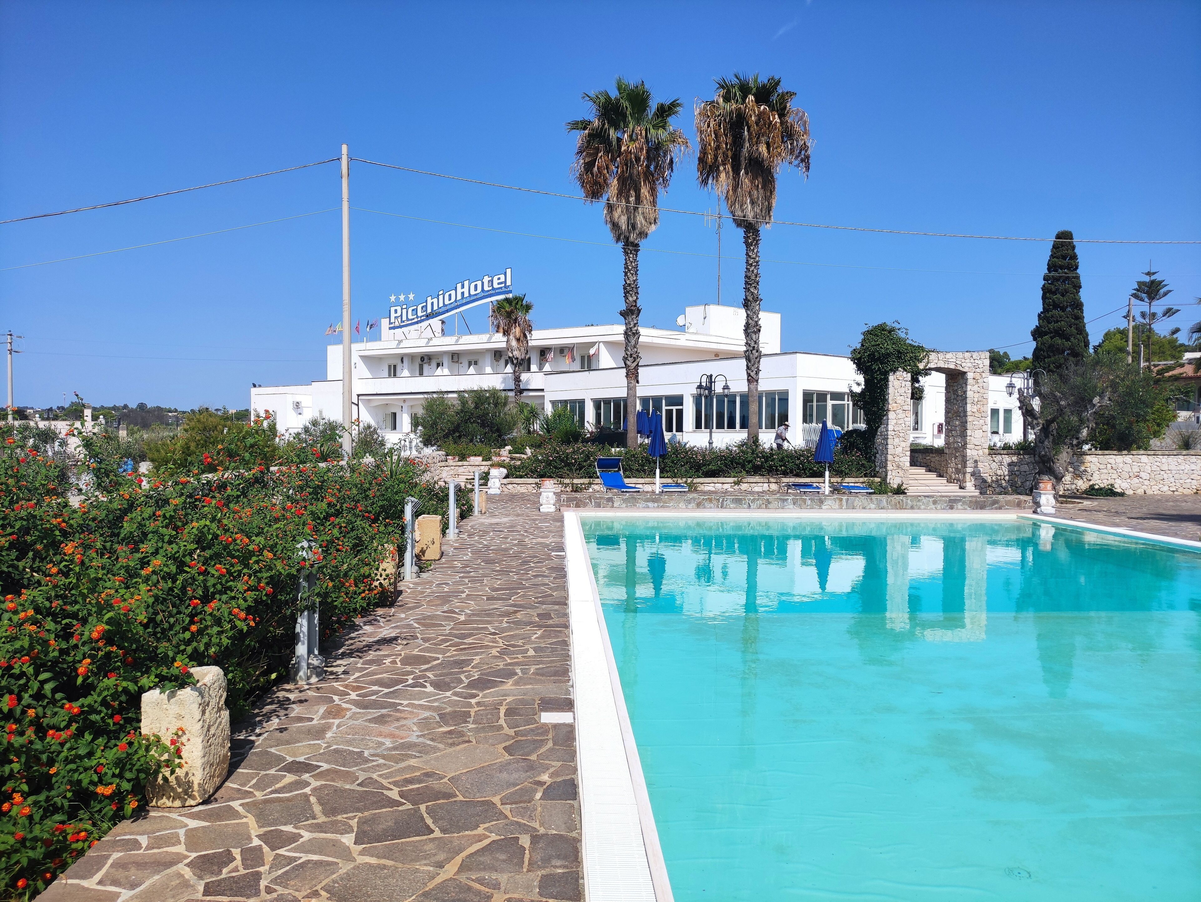Un hotel con piscina, camino de piedra, palmeras y un cielo azul despejado.