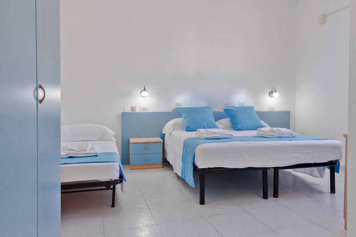 A bedroom with two beds, each with a blue headboard and white bedding. There are bedside tables with lamps and a small drawer.