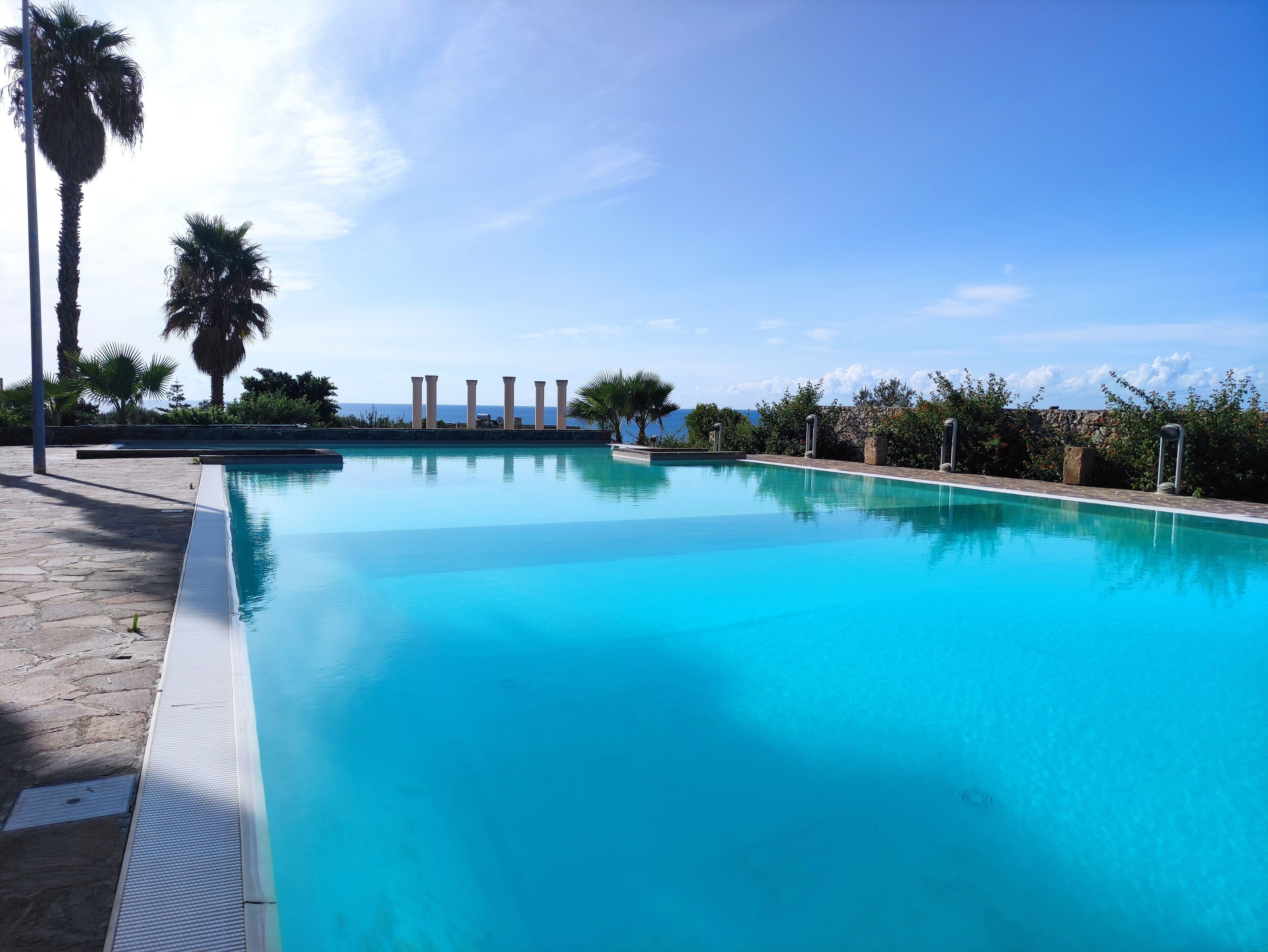 Uma piscina azul límpida com deck de pedra, palmeiras e vista para o oceano.
