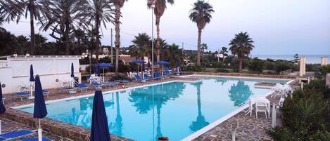 A pool area with lounge chairs and umbrellas, surrounded by palm trees and a view of the sea.