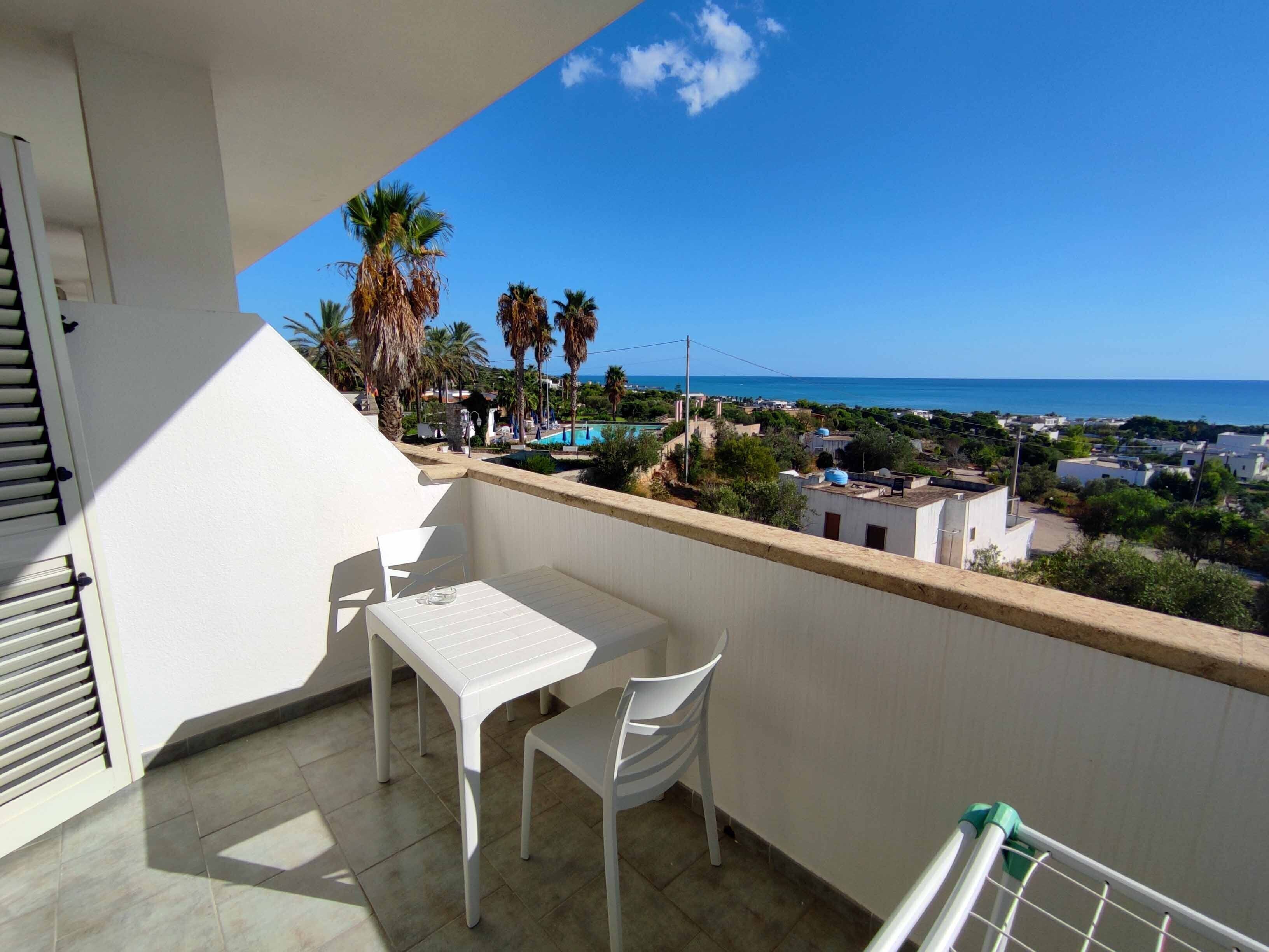 Un balcón con una mesa y sillas blancas, con vistas a un pueblo costero y al mar.