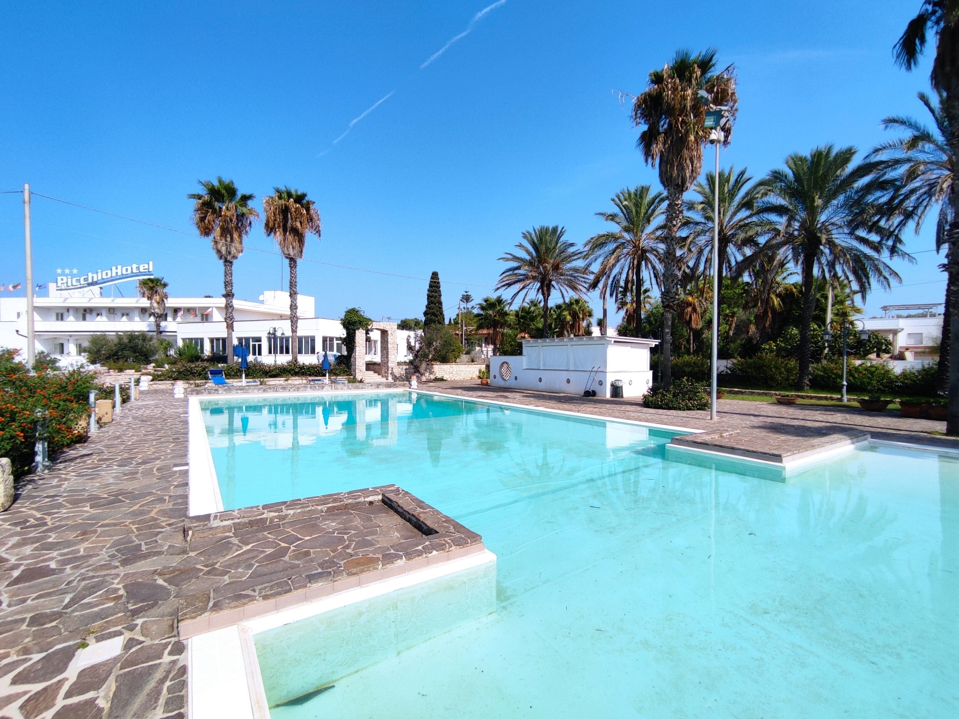 A swimming pool with stone tiles, palm trees, and a hotel building in the background.