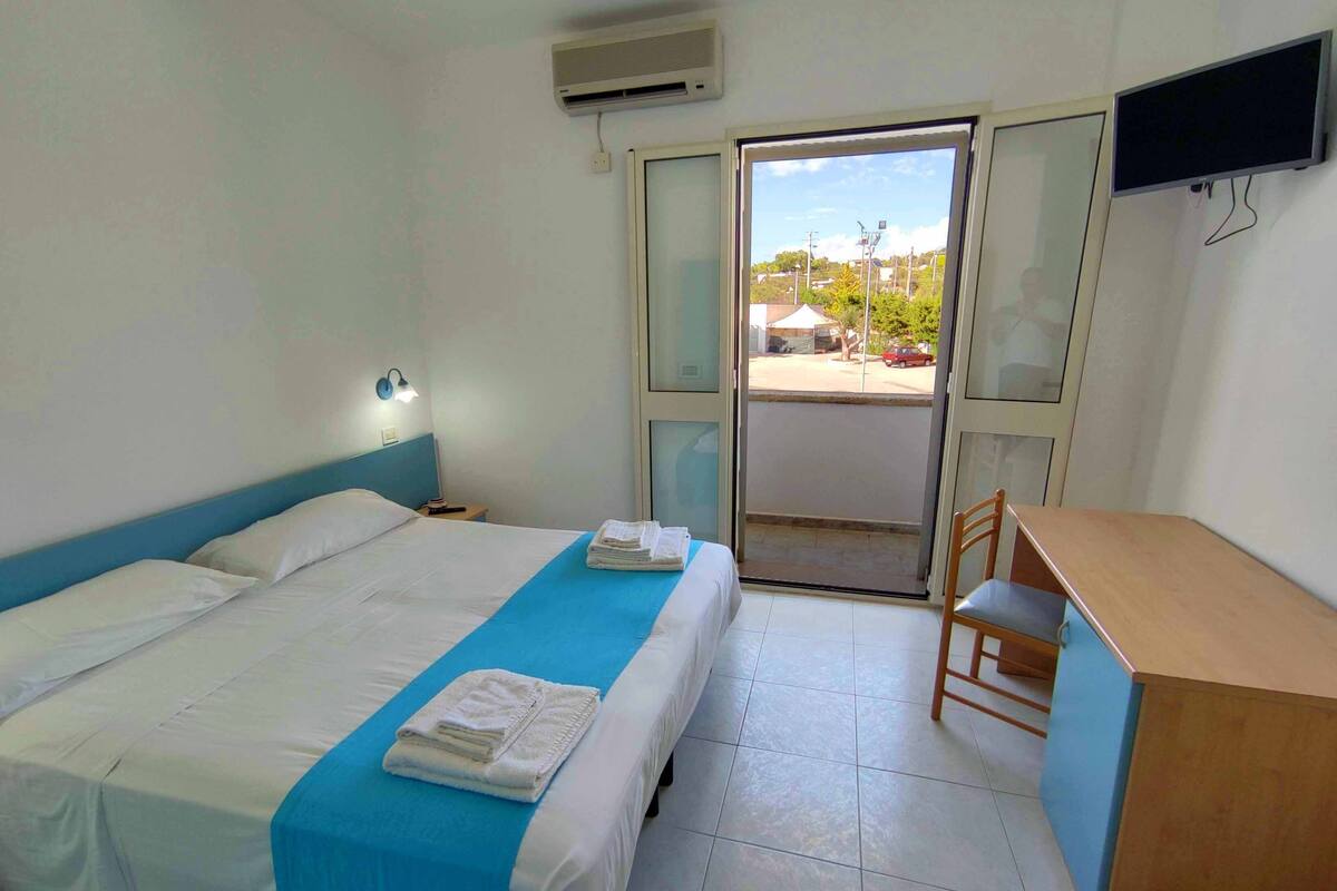 A hotel room with a bed, a desk, and a chair. There is a window with a view of the outside.
