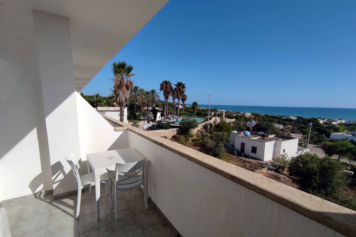 A balcony with a table and chairs, overlooking a coastal town with buildings and palm trees.