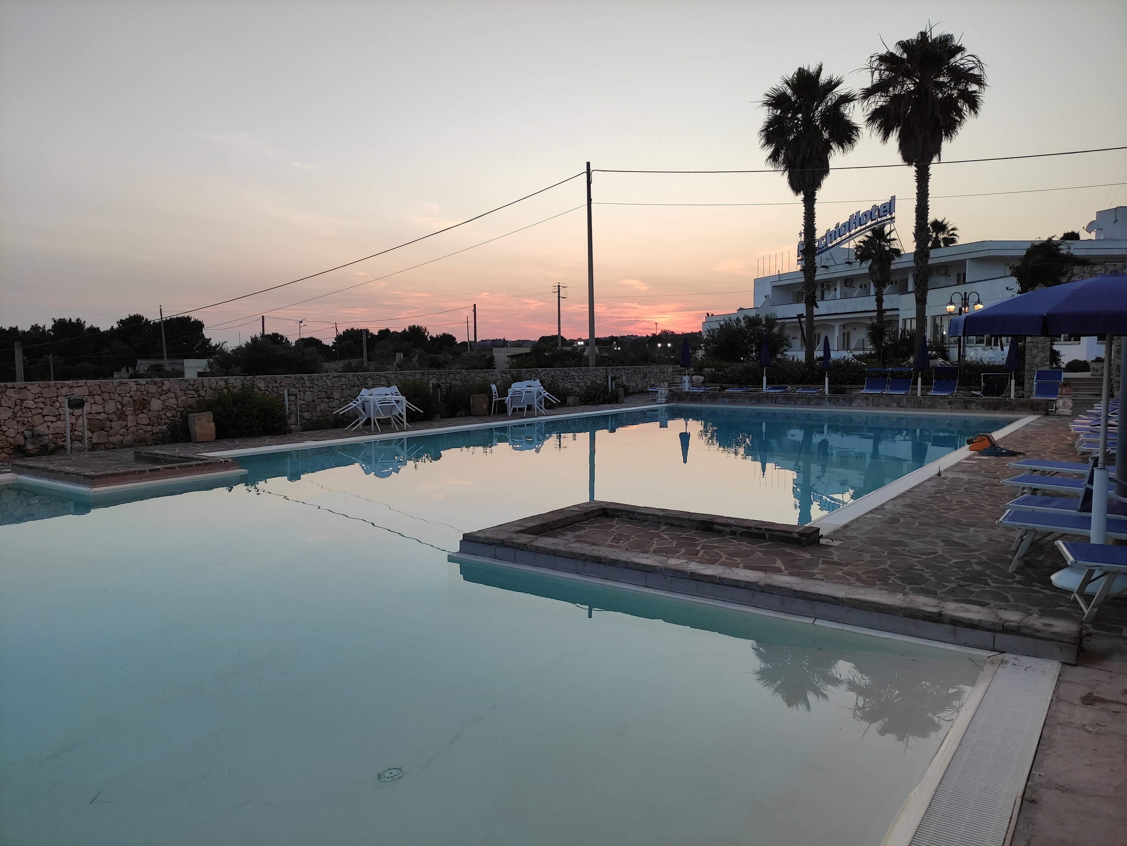 Une piscine entourée de murs de pierre et de palmiers, agrémentée de chaises longues et de parasols, avec un hôtel en arrière-plan.