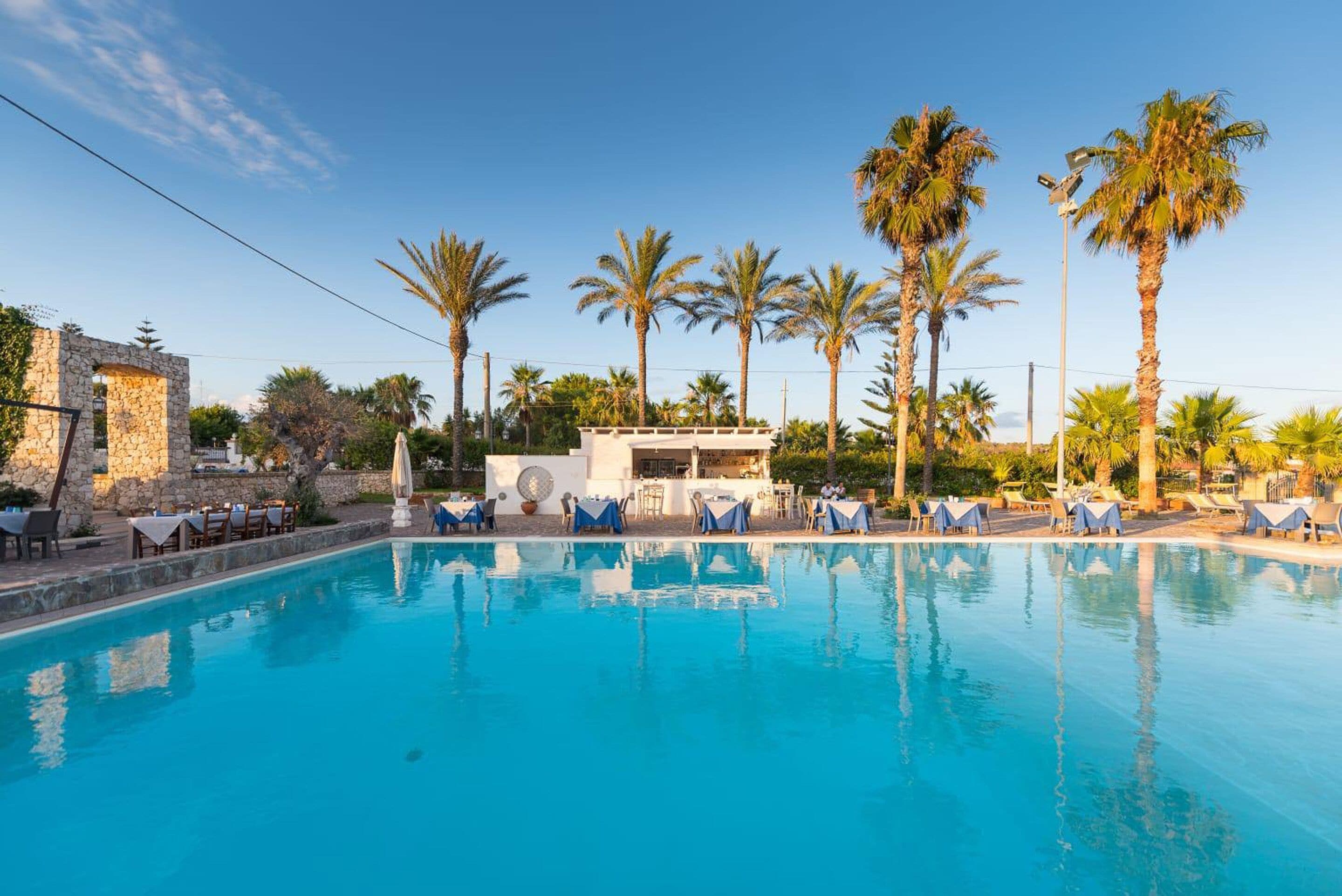 A pool area with lounge chairs and umbrellas, surrounded by palm trees and a stone building.