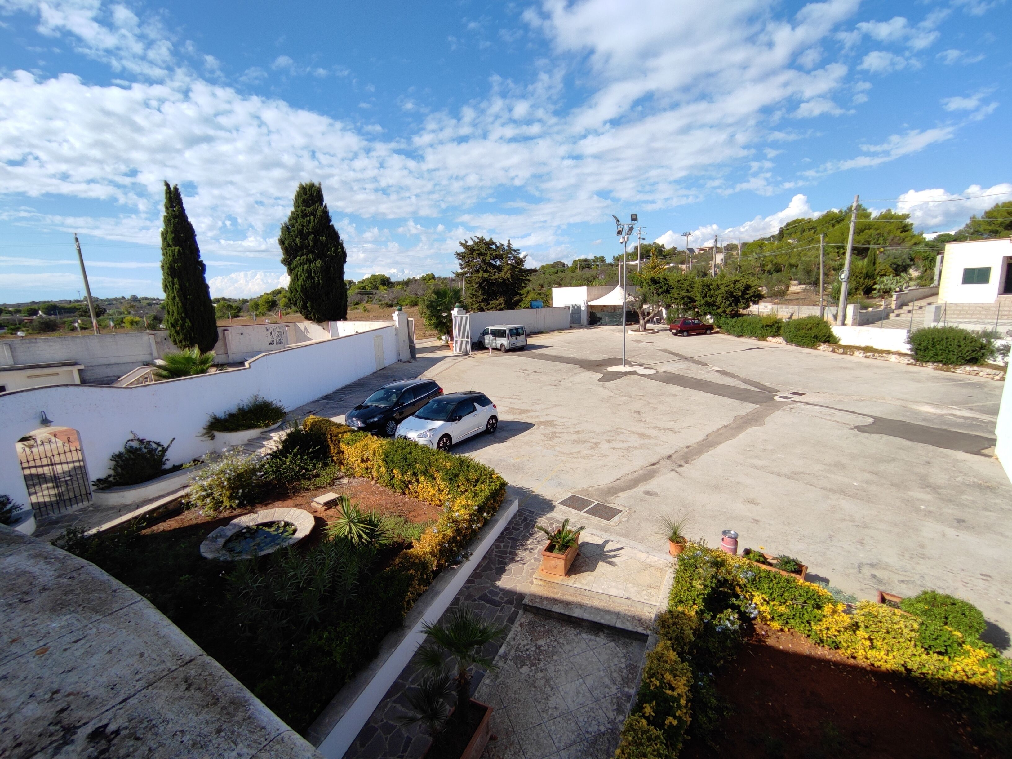 A parking area with several cars, a white building, and a garden with various plants.