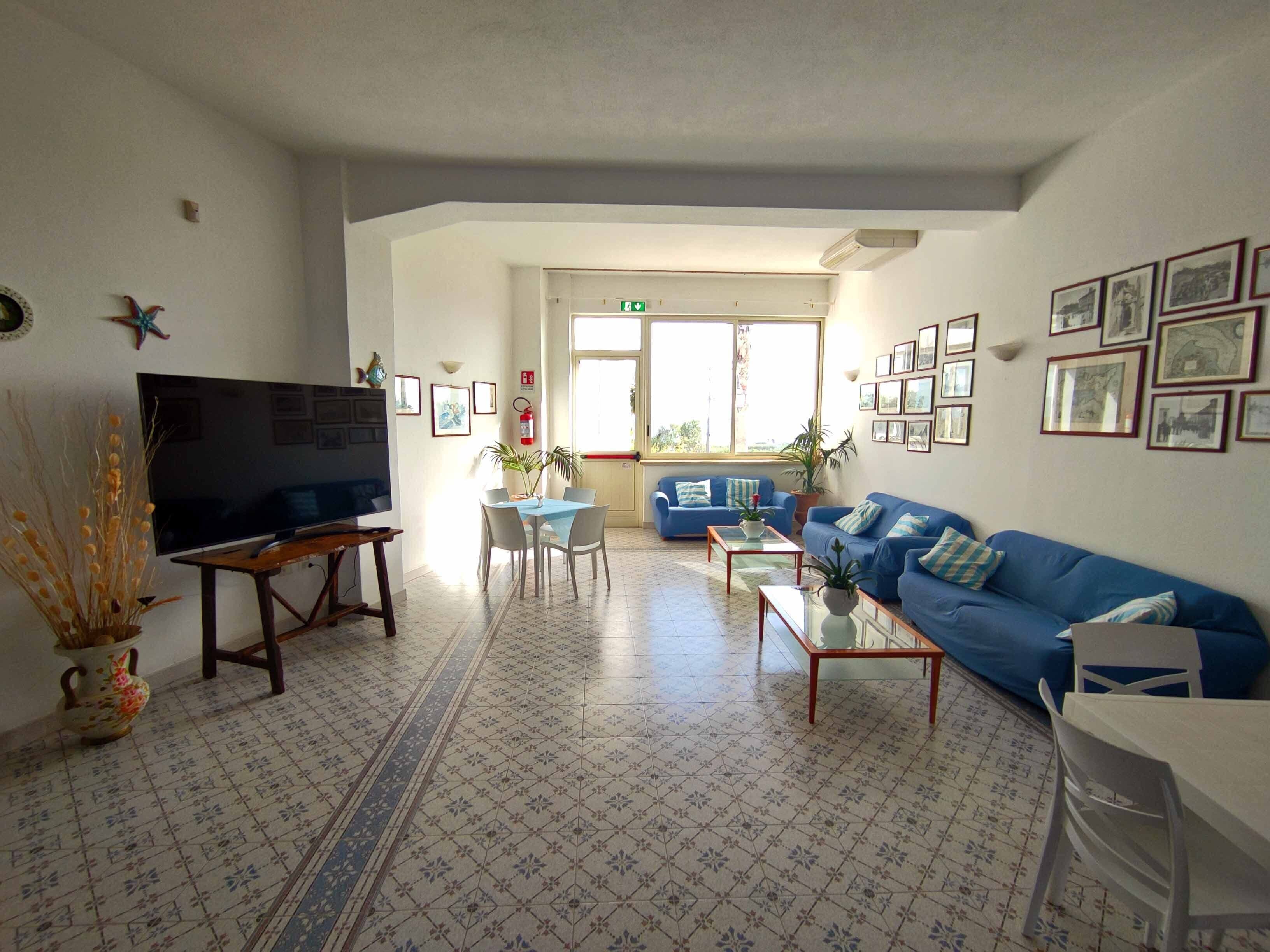 A living room with a TV, blue sofas, a dining table, and a patterned floor.