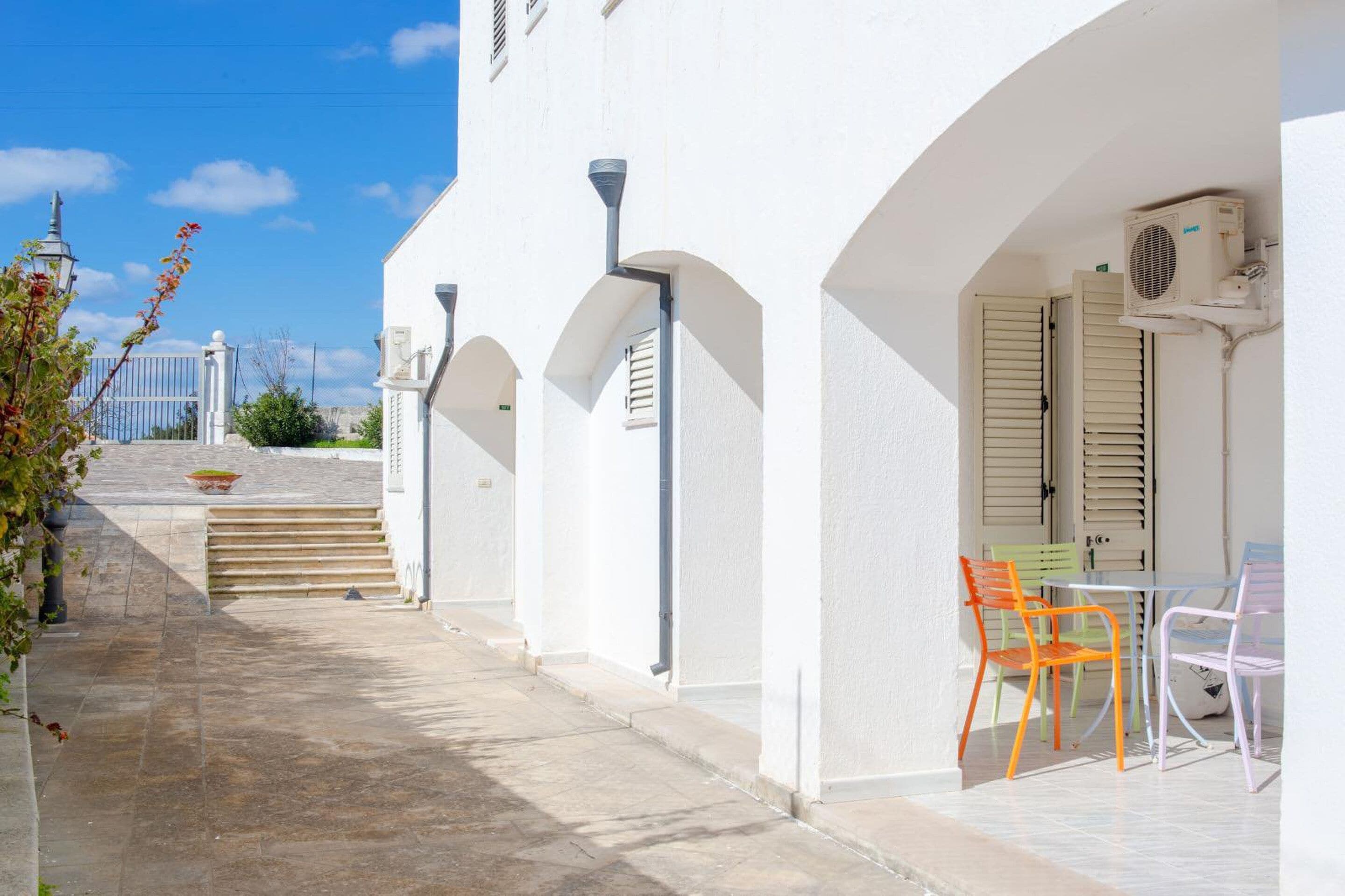 Un bâtiment blanc doté d’un espace extérieur abrité avec des sièges et d’un escalier menant à une porte.