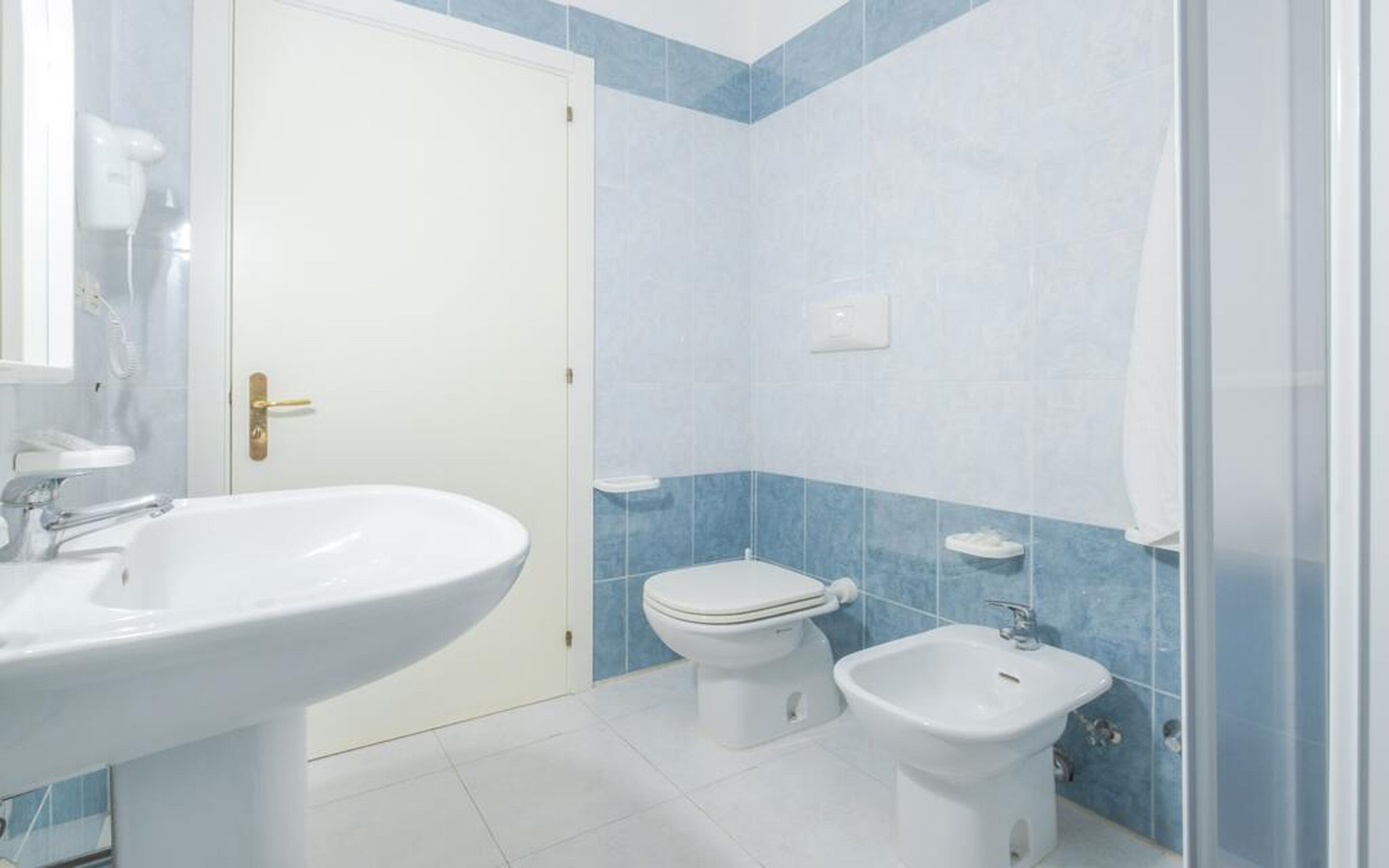Un baño con lavamanos, inodoro y bidé blancos, paredes con azulejos azules y una puerta blanca.