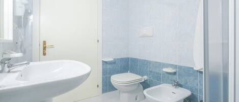 Un baño con lavamanos, inodoro y bidé blancos, paredes con azulejos azules y una puerta blanca.