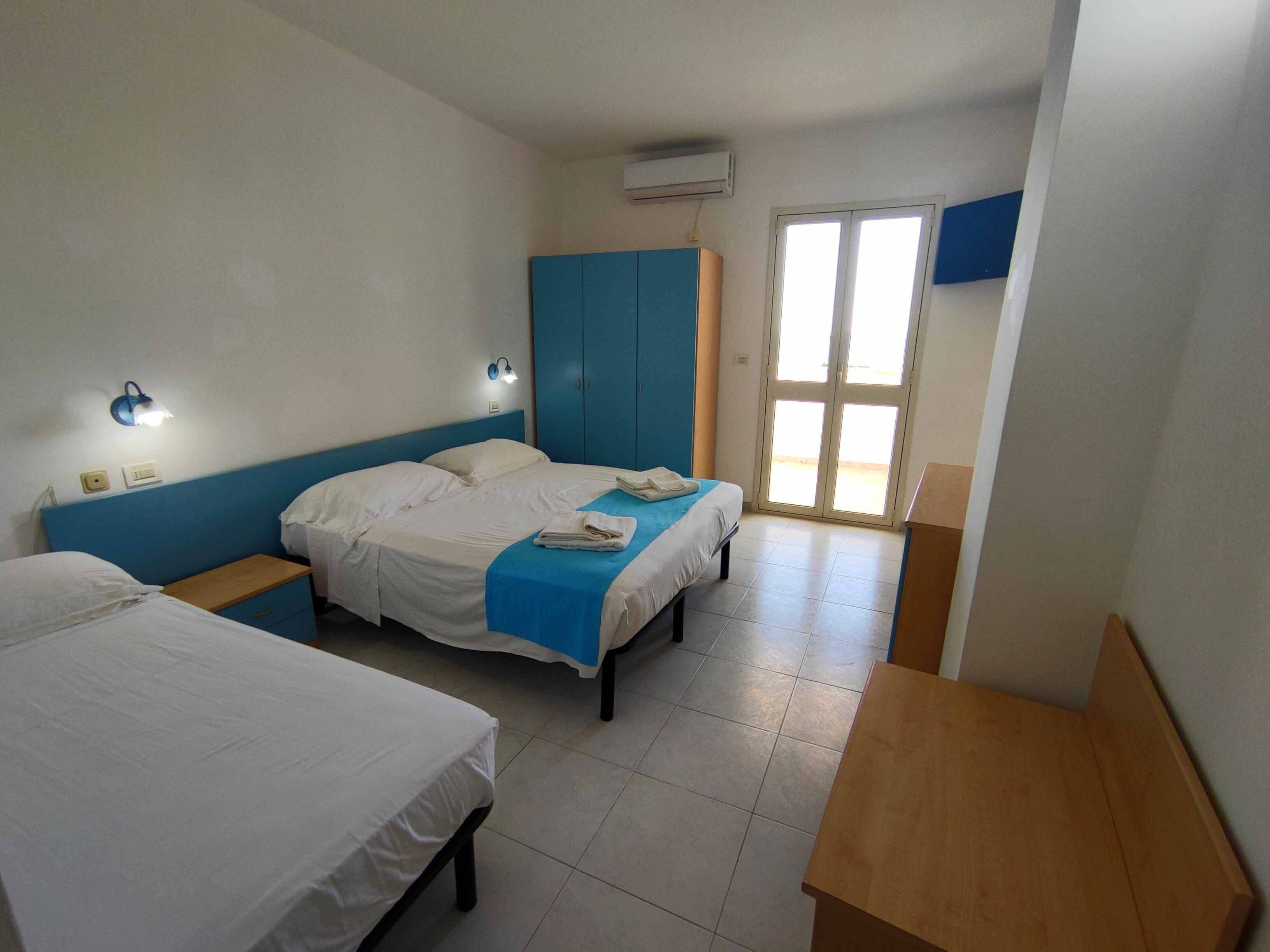 A hotel room with two beds, a blue accent wall, a wooden nightstand, and a window with a view.