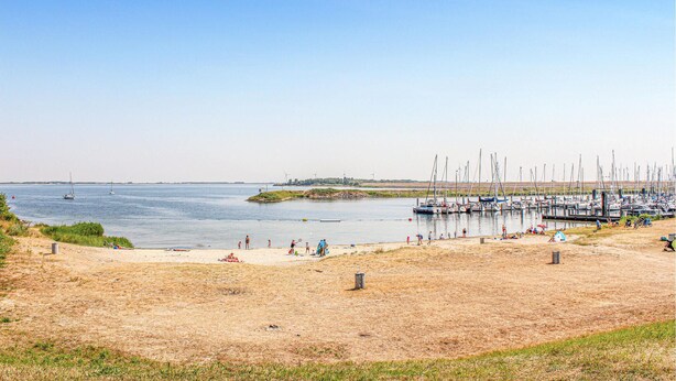 A beach with a marina filled with sailboats, a sandy shore, and a distant wind farm. - Stunning apartment in Bruinisse (Bruinisse)