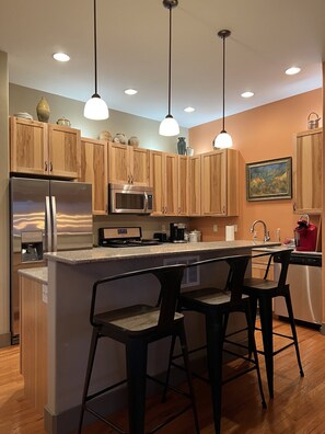 A modern kitchen with wooden cabinets, stainless steel appliances, a central island with bar stools, pendant lighting, and a painting on the wall.