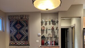 Uma área de refeições com uma mesa de madeira, jogos americanos vermelhos e uma tigela central. Há um tapete estampado na parede, uma prateleira com objetos decorativos e uma área de cozinha ao fundo.