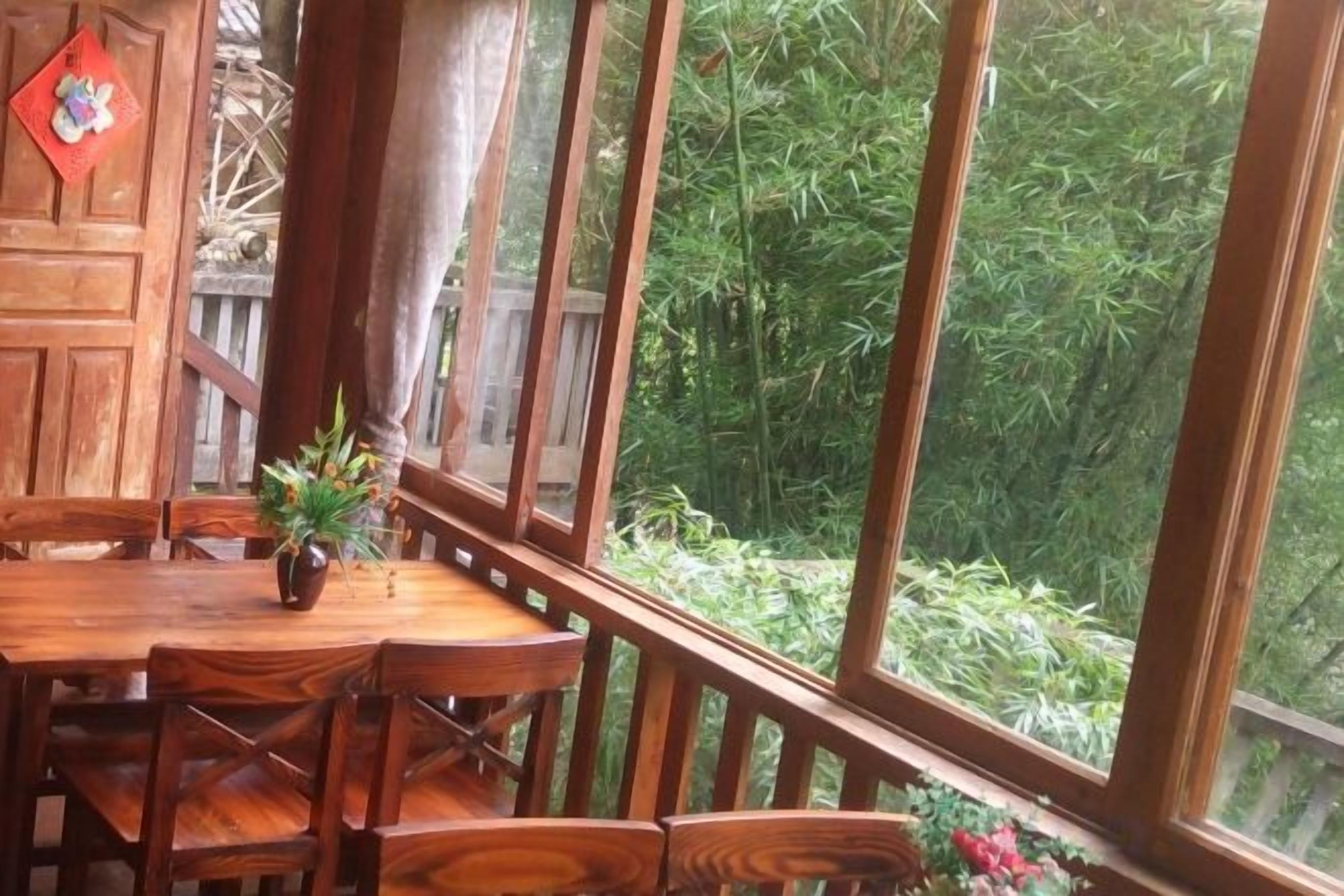 Uma mesa de jantar de madeira com cadeiras, um vaso com planta e vista para bambuzais por grandes janelas.
