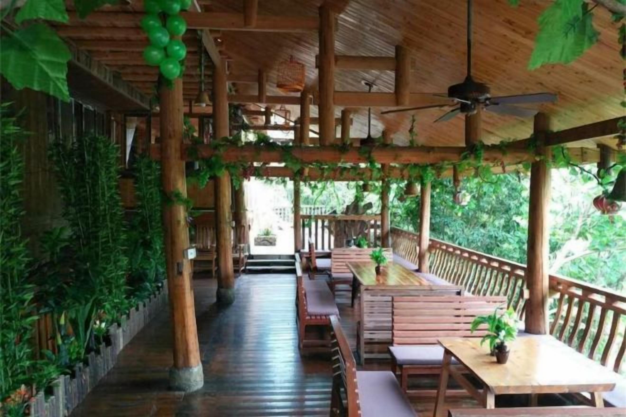 An indoor wooden structure with tables and benches, a ceiling fan, and greenery.