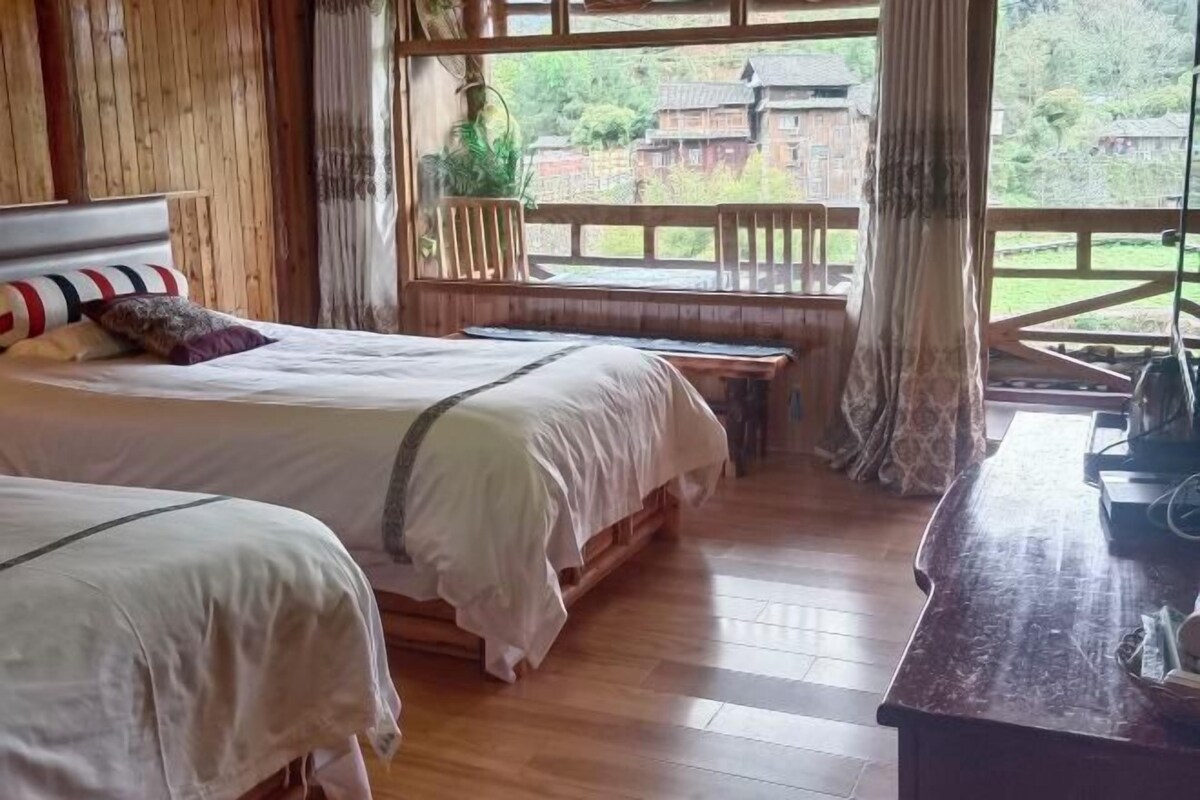 Ein Zimmer mit zwei Betten, einem hölzernen Kopfteil, einem Schreibtisch mit Computer und Blick auf eine Holzterrasse und Grünflächen.